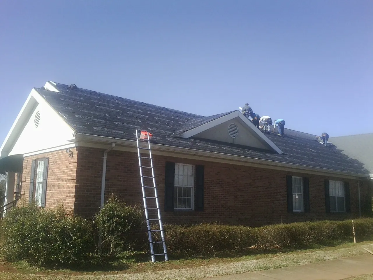 Crew tearing shingles off a brick ranch roof with a ladder set — Metal Roof Repair work by our crew in Manlius