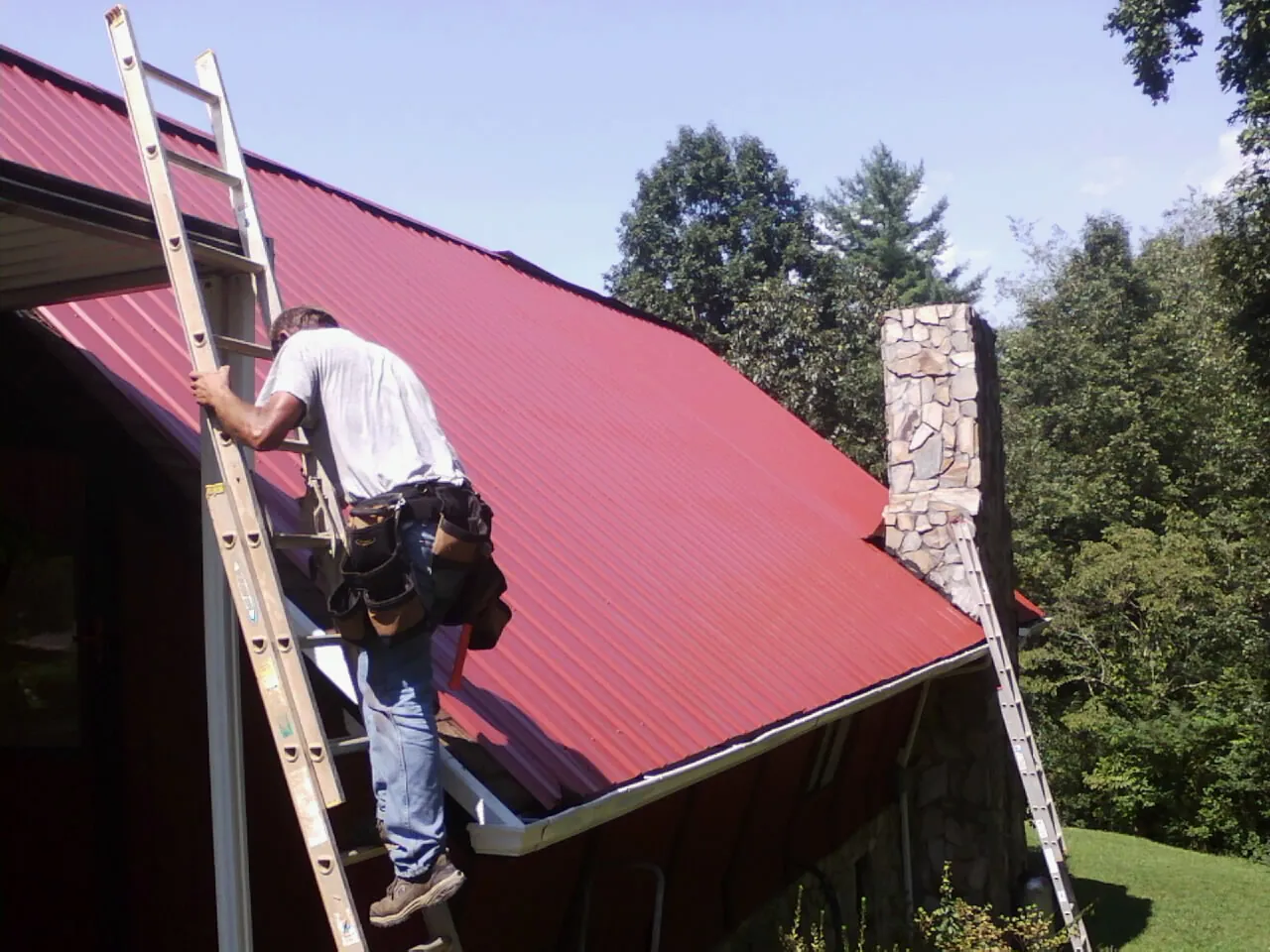 Roofer on a ladder beside a red metal roof and stone chimney — Soffit & Fascia Repair by Roofing Brothers in Manlius